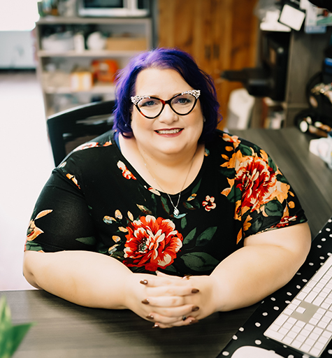 Michelle is a middle-aged, plus-sized woman with a ready smile and short purple hair. She is wearing glasses with rhinestones on them, and a black dress with orange and red roses on it.