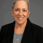 Debby Gilbert - a woman with kind approachable warm smile wearing dark jacket and a light jacket, her hair is in a neat top bun.